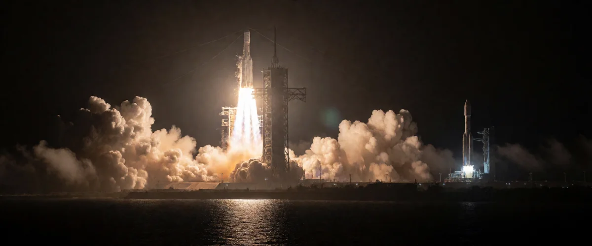 Night launch of a heavy rocket from a coastal spaceport, brilliant exhaust plume reflecting on dark ocean water and low clouds, service tower silhouetted beside the rising vehicle, a second rocket lit on a distant pad