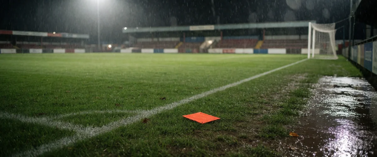 A rain-soaked football pitch at a modest ground under floodlights at night, empty terraces and wet turf glistening, a flat orange-red marker lying on the grass inside the corner arc, a white-framed goal at right, no people.
