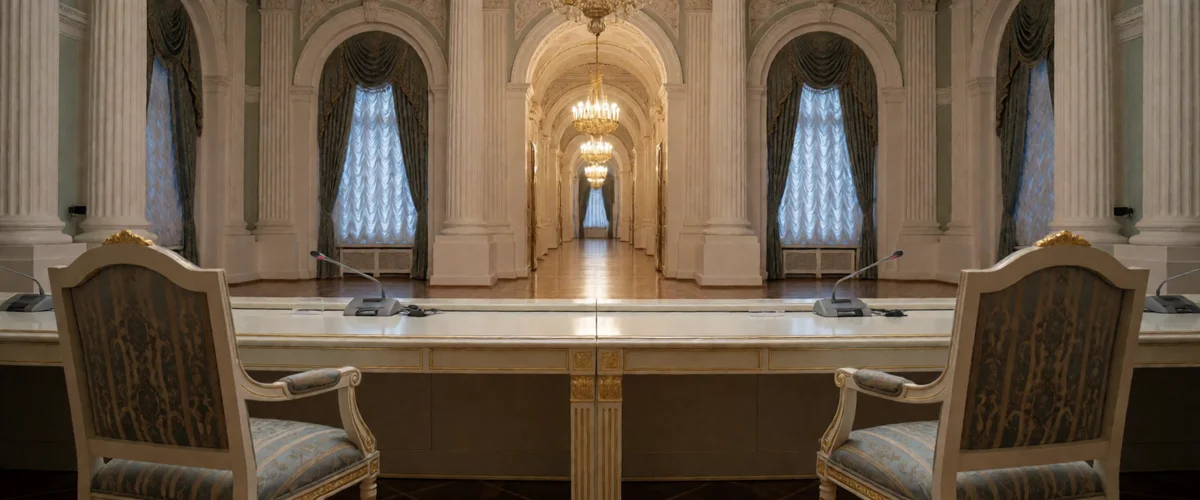An empty ornate palace conference hall with a long table fitted with gooseneck microphones and two gold-trimmed armchairs under crystal chandeliers