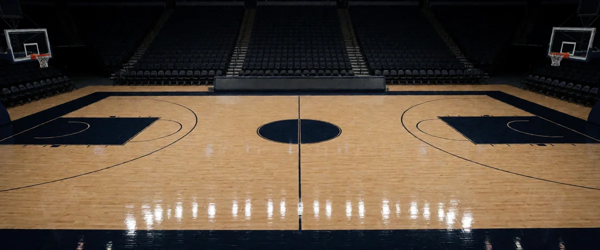 An empty basketball arena court moments before a night game, polished hardwood reflecting rows of overhead lights, center circle and painted arcs gleaming, hoops at either end, darkened stands in the background, no people.